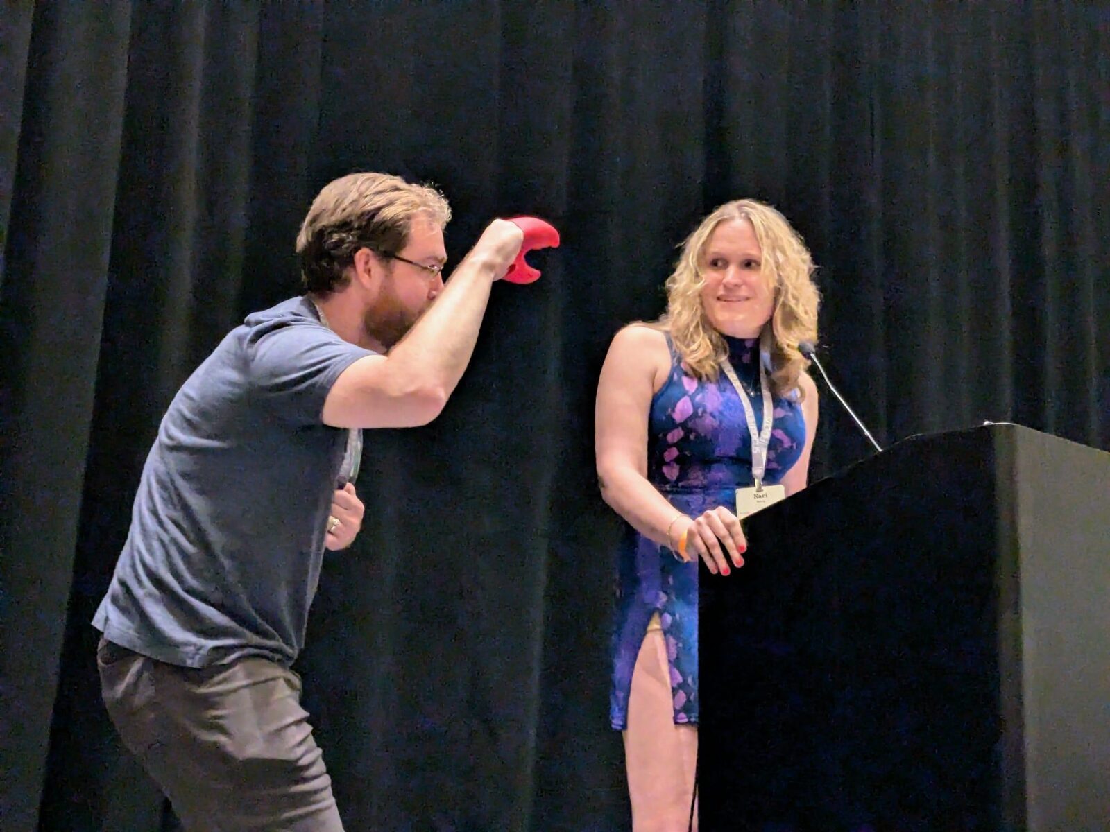 Photograph showing a man with a beard holding a red lobster claw - while deviously looking at a blonde woman at a podium who is smirking at him. She is wearing a blue top and beige pants' style='width: 90%;