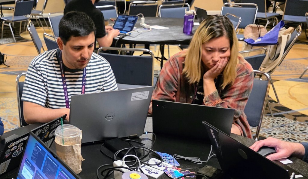 Two PyCon US 2026 sprinters sit side by side at a round table, focused on their laptops during the pyOpenSci sprint. pyOpenSci stickers, power cables, and drinks are scattered on the table while other groups work at tables across the conference hall behind them.